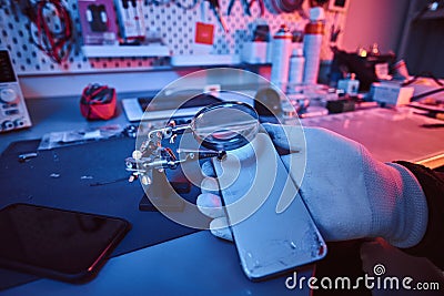 Electronic technician holds a modern smartphone with a broken body, carefully examines the damage using a magnifying Stock Photo