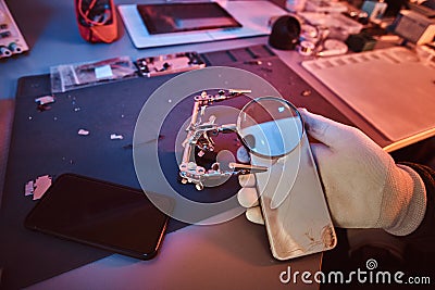 Electronic technician holds a modern smartphone with a broken body, carefully examines the damage using a magnifying Stock Photo