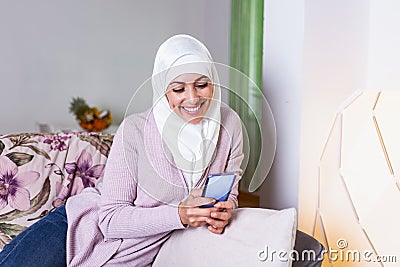 Elegant attractive female muslim using mobile smartphone searching online information when she sitting on sofa couch relaxing. Stock Photo