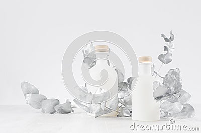 Elegant blank white glass bottles for cosmetic on soft light white wood table with silver decorative leaves as template. Stock Photo