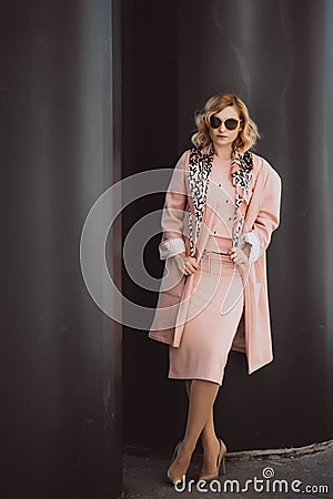 Stylish blonde girl in pink clothes and sunglasses posing in the city on a background of black huge barrels Stock Photo