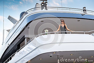 The elegant girl dressed in an evening dress of black color and sunglasses stands on the top deck of a huge yacht in Stock Photo