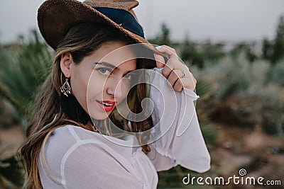Elegant long-haired girl in stylish hat looks back, while walking in beautiful exotic park. Close-up portrait of pretty Stock Photo