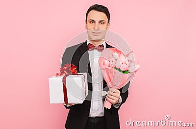Elegant man, in a suit, with a bouquet of flowers, and a gift box, on a pink background, the concept of women`s day Stock Photo