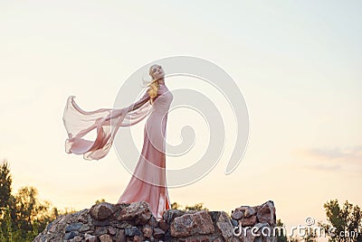 Elegant princess with blond fair wavy hair with tiara on it, wearing a long light pink rose fluttering dress, standing Stock Photo