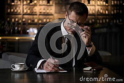 An elegant,serious and concentrated businessman sitting in modern restaurant and making the notes in his notebook Stock Photo