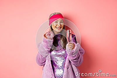 Elegant and stylish asian woman in purple faux fur coat eating donuts and drinking champagne, having fun at party Stock Photo