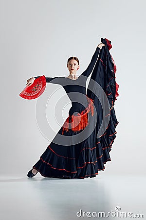 Elegant woman, flamenco dancer with fan in hand, black and red dress performing against grey studio background Stock Photo