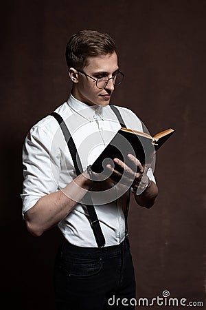 A man in round glasses, suspenders, a white shirt is reading a book Stock Photo