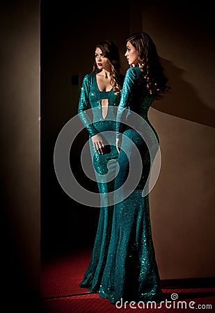 Elegant young woman in turquoise long dress looking into a large mirror, side view. Beautiful slim girl with creative hairstyle Stock Photo