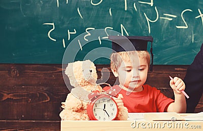 Elementary education concept. First former with toy on desk. Pupil in mortarboard, chalkboard on background. Boy on busy Stock Photo