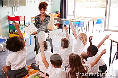 Elementary Pupils Wearing Uniform Raise Hands To Answer Question As Female Teacher Reads Book Stock Photo