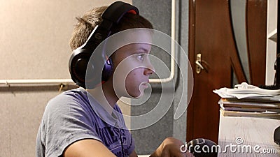 an eleven-year-old boy in headphones sits at the computer in the evening and plays games and studies Stock Photo
