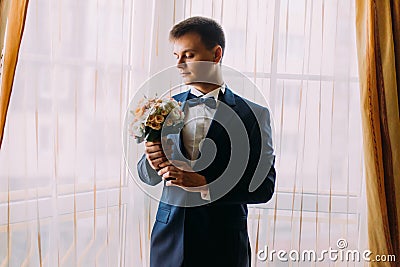 Ellegant dressed romantic groom holding a wedding bouquet of pale pink roses in hands standing near the window Stock Photo