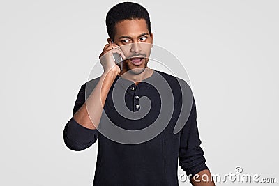 Embarrassed African American man has telephone coversation indoor, discusses something unpleasant and surprising, looks Stock Photo