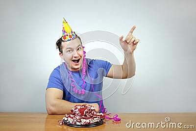 Emotional beautiful caucasian man with birthday party cone hat on head and crumple cake raises your index finger up, smiling happi Stock Photo