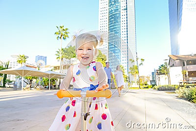 Emotional cute little blondy toddler girl in dress riding scooter in the city park recreation area with modern skyscraper building Stock Photo