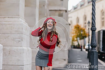 Emotional female model in soft red sweater posing in front of old white building. Portrait of graceful european girl Stock Photo