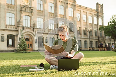 Emotional guy sitting with books and a laptop on a lawn on the background of a university building,reading a notebook with Stock Photo