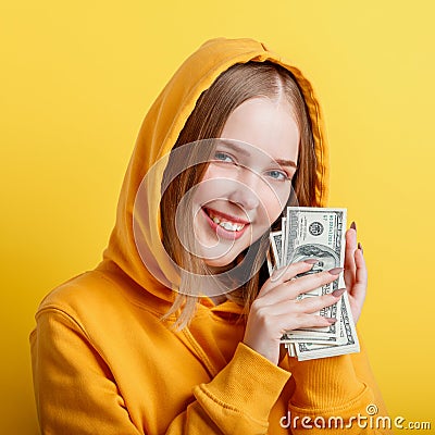 Emotional happy teenage blonde girl in hood win money cash holding dollars in hands isolated on color yellow background. Portrait Stock Photo