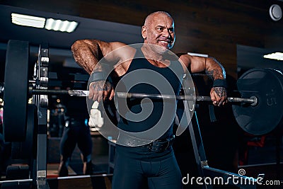 Emotional Older bald bodybuilder doing trapezius and deltoid exercise with a barbell in the gym Stock Photo