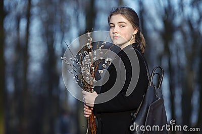 Emotional portrait of young happy beautiful woman with a bouquet of pussy-willows wearing black coat strolling at evening golden h Stock Photo