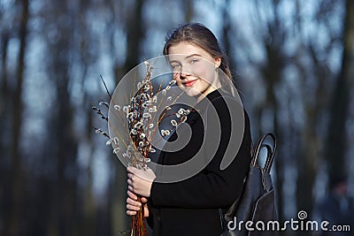 Emotional portrait of young happy beautiful woman with a bouquet of pussy-willows wearing black coat strolling at evening golden h Stock Photo