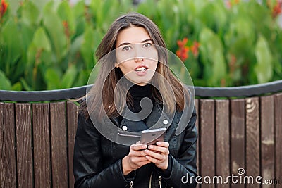 Emotional portrait of a young pretty frustrated brunette woman sitting outdoors city park wearing black leather coat using smartph Stock Photo