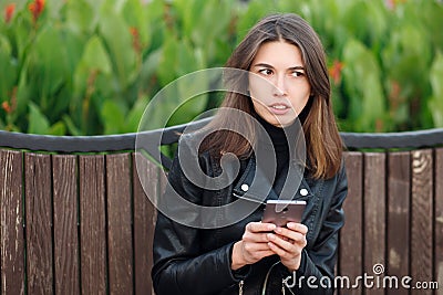 Emotional portrait of a young pretty frustrated brunette woman sitting outdoors city park wearing black leather coat using smartph Stock Photo