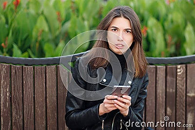 Emotional portrait of a young pretty frustrated brunette woman sitting outdoors city park wearing black leather coat using smartph Stock Photo