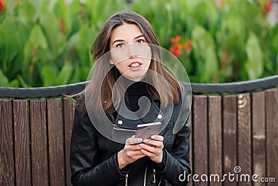 Emotional portrait of a young pretty frustrated brunette woman sitting outdoors city park wearing black leather coat using smartph Stock Photo