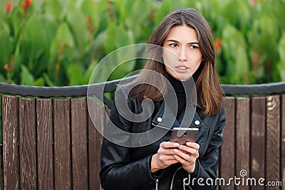 Emotional portrait of a young pretty frustrated brunette woman sitting outdoors city park wearing black leather coat using smartph Stock Photo