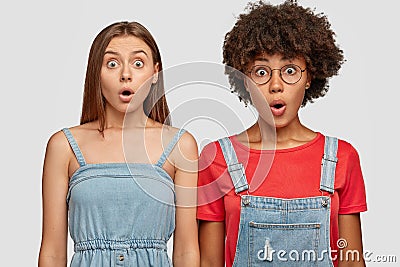 Emotional stupefied mixed race women stand closely to each other, stare with bugged eyes, dressed in casual clothes Stock Photo