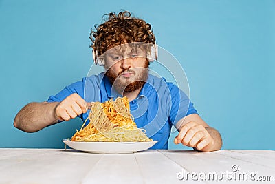 Emotional young hairy red-bearded man tasting large portion of noodles, pasta isolated on blue studio background. Stock Photo