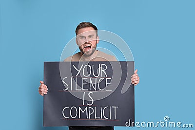 Emotional young man holding sign with phrase Your Silence Is Complicit on light blue background. End racism Stock Photo