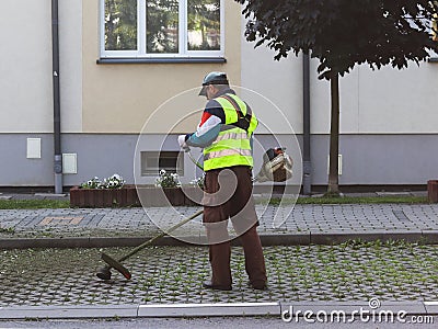 An employee of the municipal service of the city removes the territory. Refinement of the area around the house. Mowing of grass b Editorial Stock Photo