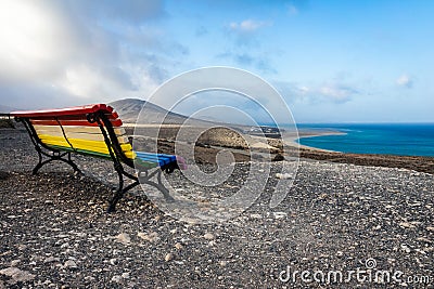 An empty bench, painted in a rainbow of colors and a view of Playa de Sotavento de Jandia. Stock Photo