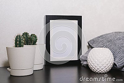 An empty black frame, cacti and succulents in pots, a candle holder and a gray sweater on a table against a white wall Stock Photo