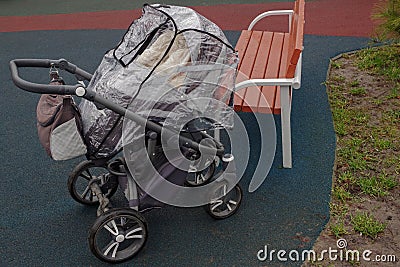 Empty four-wheeled black pram covered with anti-rain cellophane canopy, standing next to brown bench on playground on rainy day Stock Photo