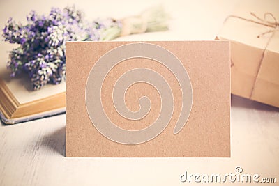 Empty greeting kraft card in front of a lavender bouquet, wrapped gift and old book over a white wood background. Vintage style. Stock Photo