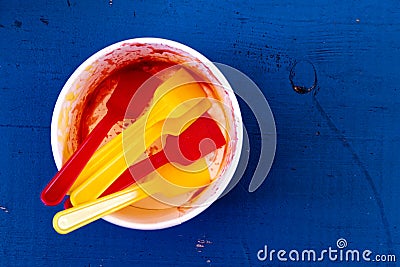 Empty messy ice cream paper cup with yellow and red plastic spoons isolated on blue painted wood from above. Stock Photo