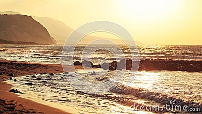 Empty wild beach lit by golden sunset. Small waves over rock, hills in hazy background Stock Photo