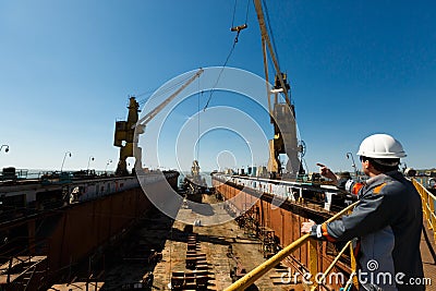 Engineer oversees vessel maintenance in dry dock. Maritime worker points, directs crane at shipyard under sunny sky Stock Photo