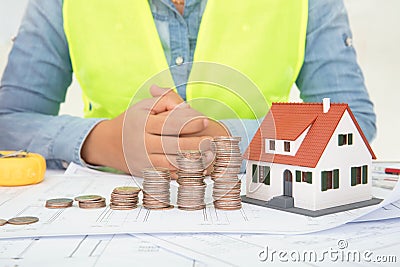 The engineer sits in front of a table with drawings and a small house model and a row of dollar coins Stock Photo