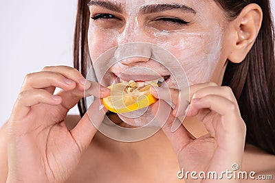 Enlarged photo of brunette woman with moisturizing facial mask bites slice of lemon. Girl with cream citrus mask on face Stock Photo