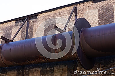 Enormous industrial pipe running along the exterior of a brick manufacturing warehouse, rusted metal and huge flanges riveted toge Stock Photo