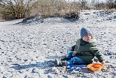 Enthusiast kid sits on a beach playing with sand. Fervid or fervent child dressed against the cold is sitting on a strand Stock Photo