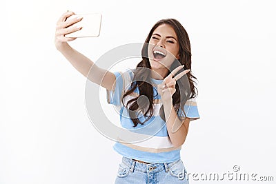 Enthusiastic happy woman posing near white background raise hand hold smartphone, taking positive cute selfie wink Stock Photo