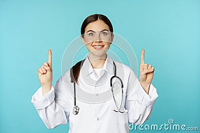 Enthusiastic medical worker, young woman doctor in white coat, stethoscope, showing advertisement, pointing fingers up Stock Photo