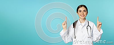 Enthusiastic medical worker, young woman doctor in white coat, stethoscope, showing advertisement, pointing fingers up Stock Photo